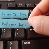 Hand placing handwritten "Return to Office" and "Welcome Back" notes in keyboard keys